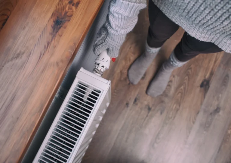 Person wearing gloves and a sweater adjusts the thermostat knob on a white radiator heater, standing on a wooden floor.