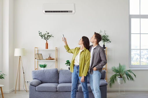 A man and a woman in a home standing in a living room. The woman is using a remote to turn on a ductless AC unit on the wall.