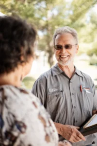 A man in a gray work shirt with a name tag reading "John" smiles while discussing a maintenance plan with a woman outdoors.