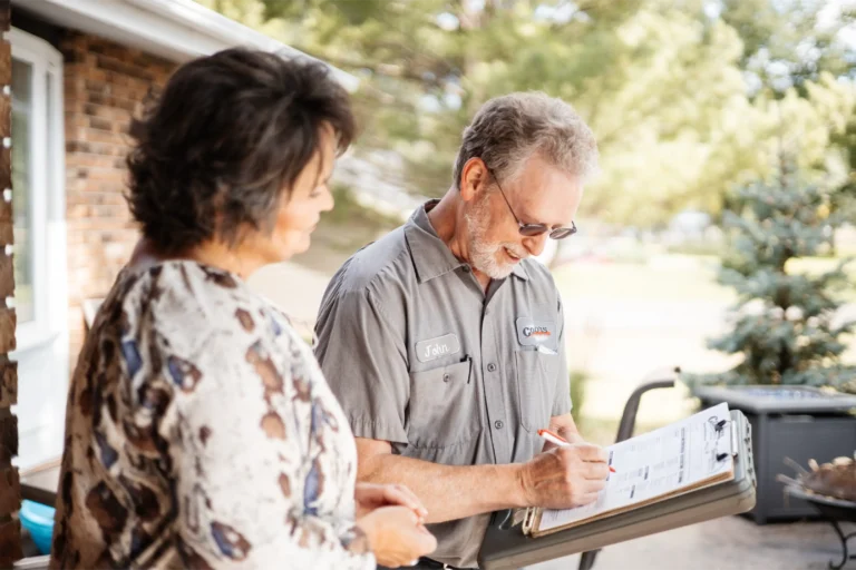 A man in a work shirt fills out a contact form on a clipboard while standing next to a woman outside a house.
