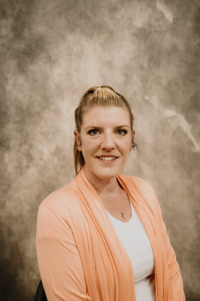 A friendly Connors team member with blonde hair in a ponytail smiles warmly, wearing a peach cardigan over a white top.