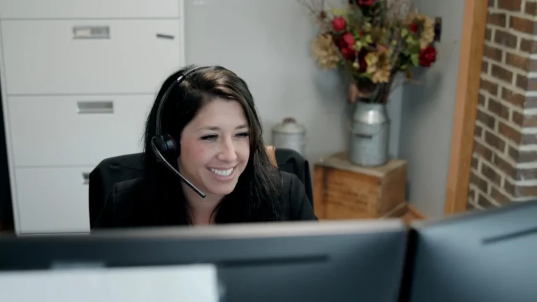 A woman with a headset sits at a desk with two monitors, smiling as she handles contact inquiries. A filing cabinet and a decorative flower arrangement are in the background.