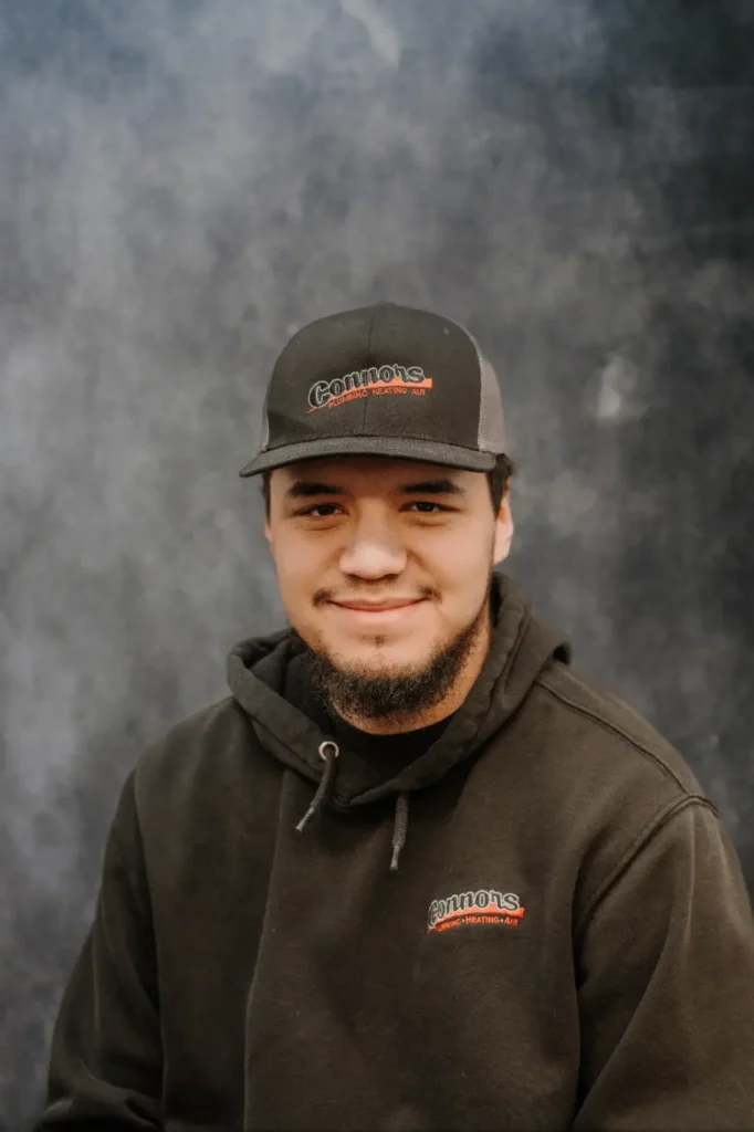 A friendly Connors Plumbing, Heating, Air, & Electrical team member smiles at the camera in branded attire against a gray backdrop.