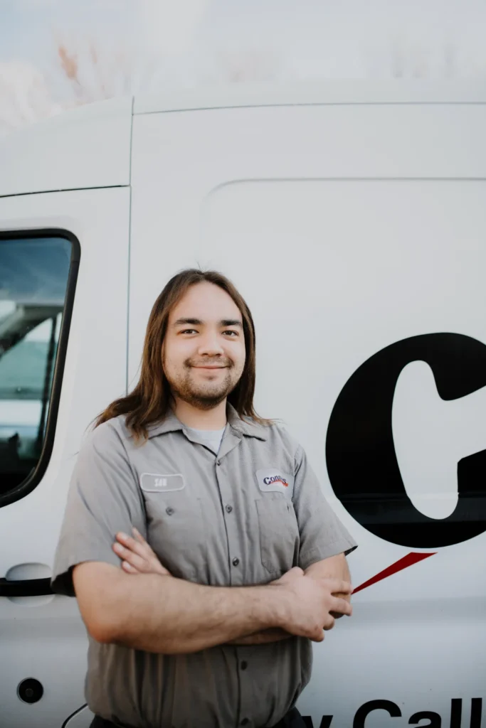 A Connors team member with long hair and a work shirt stands confidently by a service van, ready to assist customers.