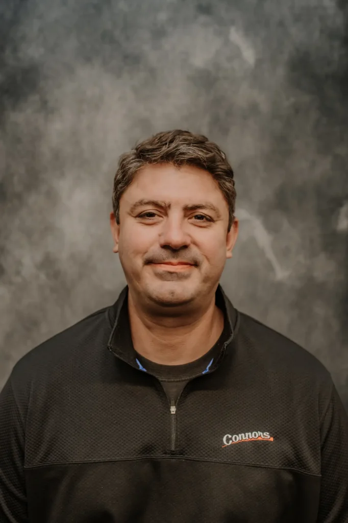 A Connors Plumbing, Heating, Air, & Electrical team member with short brown hair smiles warmly in front of a gray textured backdrop.