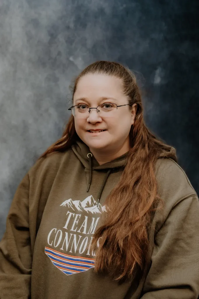 A member of the Connors team with long brown hair and glasses wears a "Team Connors" hoodie, smiling confidently in front of a dark backdrop.