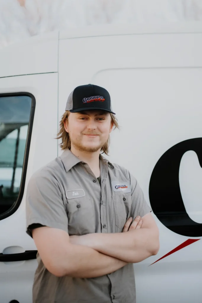A man wearing a gray work shirt and black cap stands with arms crossed in front of a white company van, ready to represent our team. Learn more about us and our dedication to quality service.
