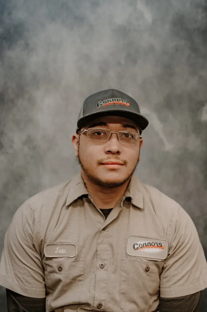 Juan, a Connors Plumbing, Heating, Air, & Electrical team member, smiles in uniform and glasses against a neutral grey background.