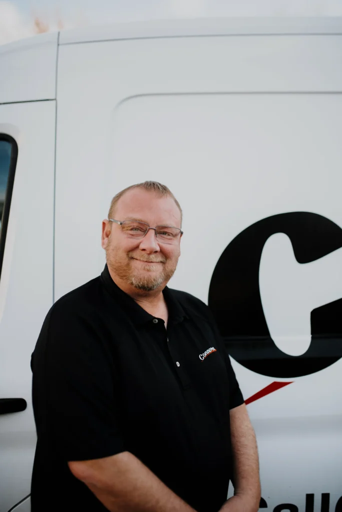 A Connors Plumbing, Heating, Air, & Electrical team member in glasses and uniform stands by a branded service van with "about us" signage.