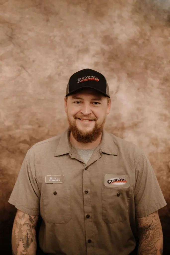 A friendly, bearded team member from Connors Plumbing, Heating, Air & Electrical stands smiling in uniform before a brown backdrop.