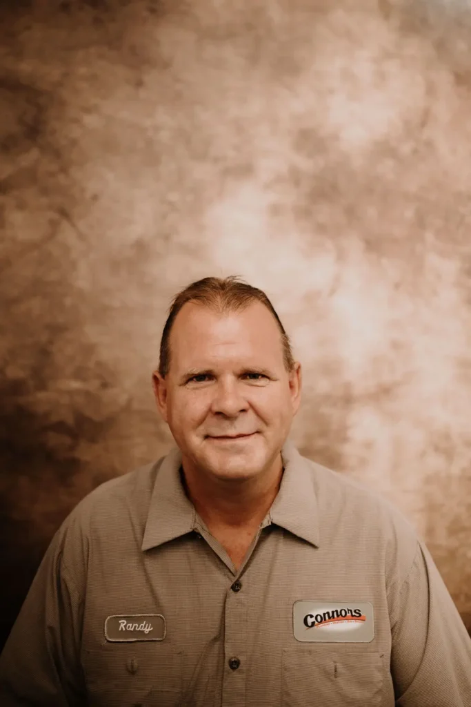 Randy, a Connors team member in a gray work shirt, smiles proudly in front of a brown backdrop for our About Us page.