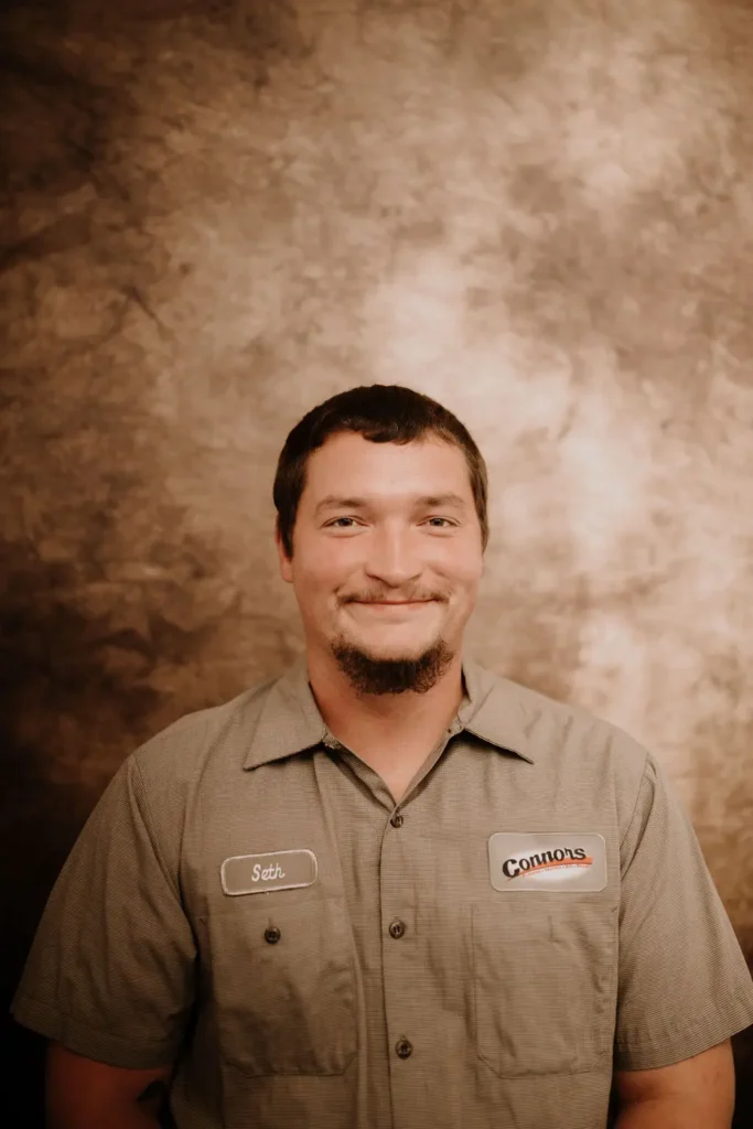 Seth from Connors Plumbing, Heating, Air, & Electrical smiles in uniform for our About Us page against a brown backdrop.