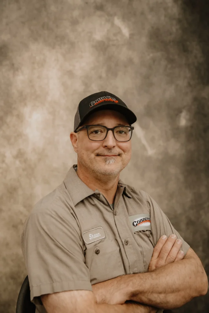 A friendly Connors team member in glasses, black cap, and gray work shirt smiles confidently in front of a brown background.