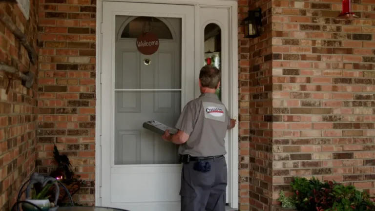 A delivery worker in uniform stands at the front door of a brick house, holding a clipboard and facing the door with a "Welcome" sign, ready to assist with 24/7 emergency service.