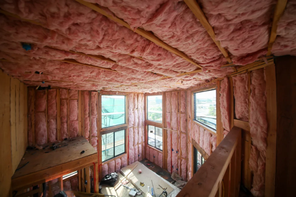 A bright, spacious room is in the midst of construction by the Connors Plumbing, Heating, Air, & Electrical team. Exposed wooden framing and pink fiberglass insulation are visible on the walls and ceiling, while large windows let in plenty of natural light as our crew works to create a comfortable living space for our valued customers.